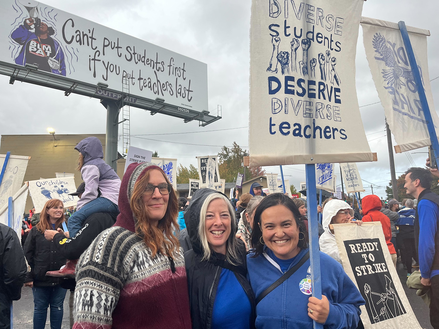 Three teachers standing in a a group of teachers in front of a sign that reads "Diverse Students deserve diverse teachers"
